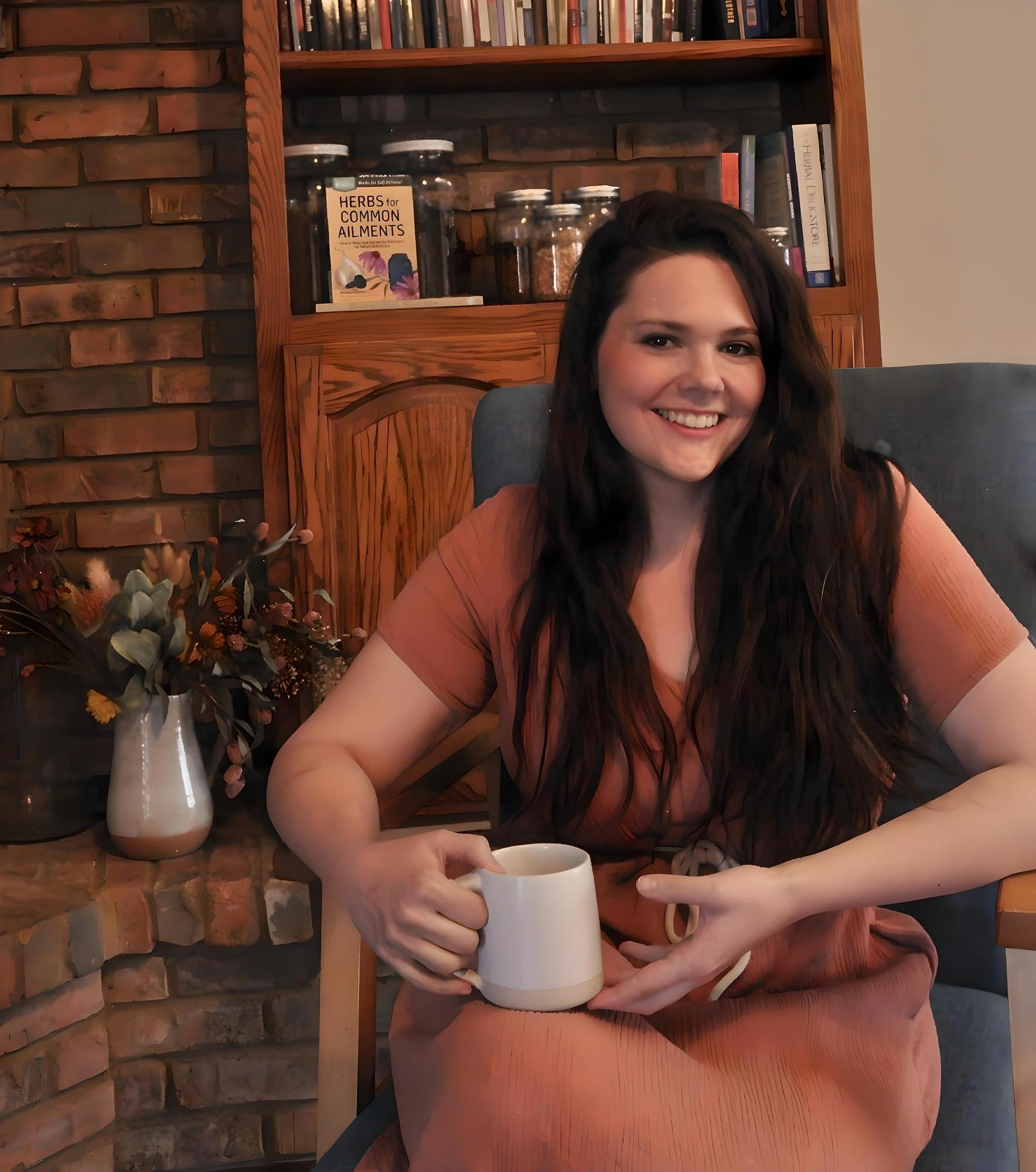 Morgan Murphy, owner of Heritage Remedy Co., sitting by her brick fireplace with a mug, shelves of herb books and jars behind her